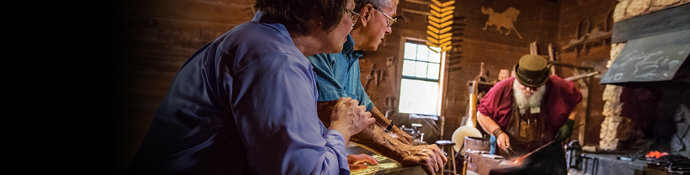 Pareja de personas mayores observa a un herrero con un martillo y un trozo de metal en el Sitio Histórico en Grand Detour, Illinois, Estados Unidos