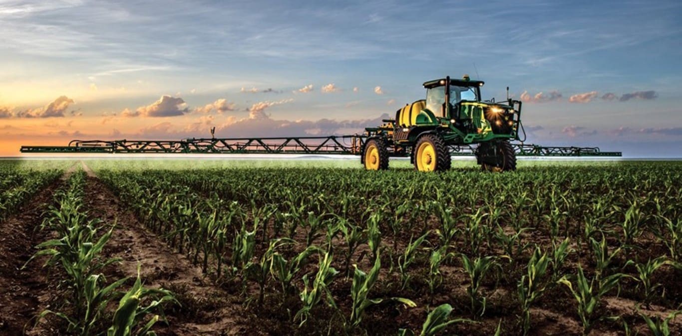 Pulverizadora John Deere trabajando en un campo de maíz con el atardecer de fondo