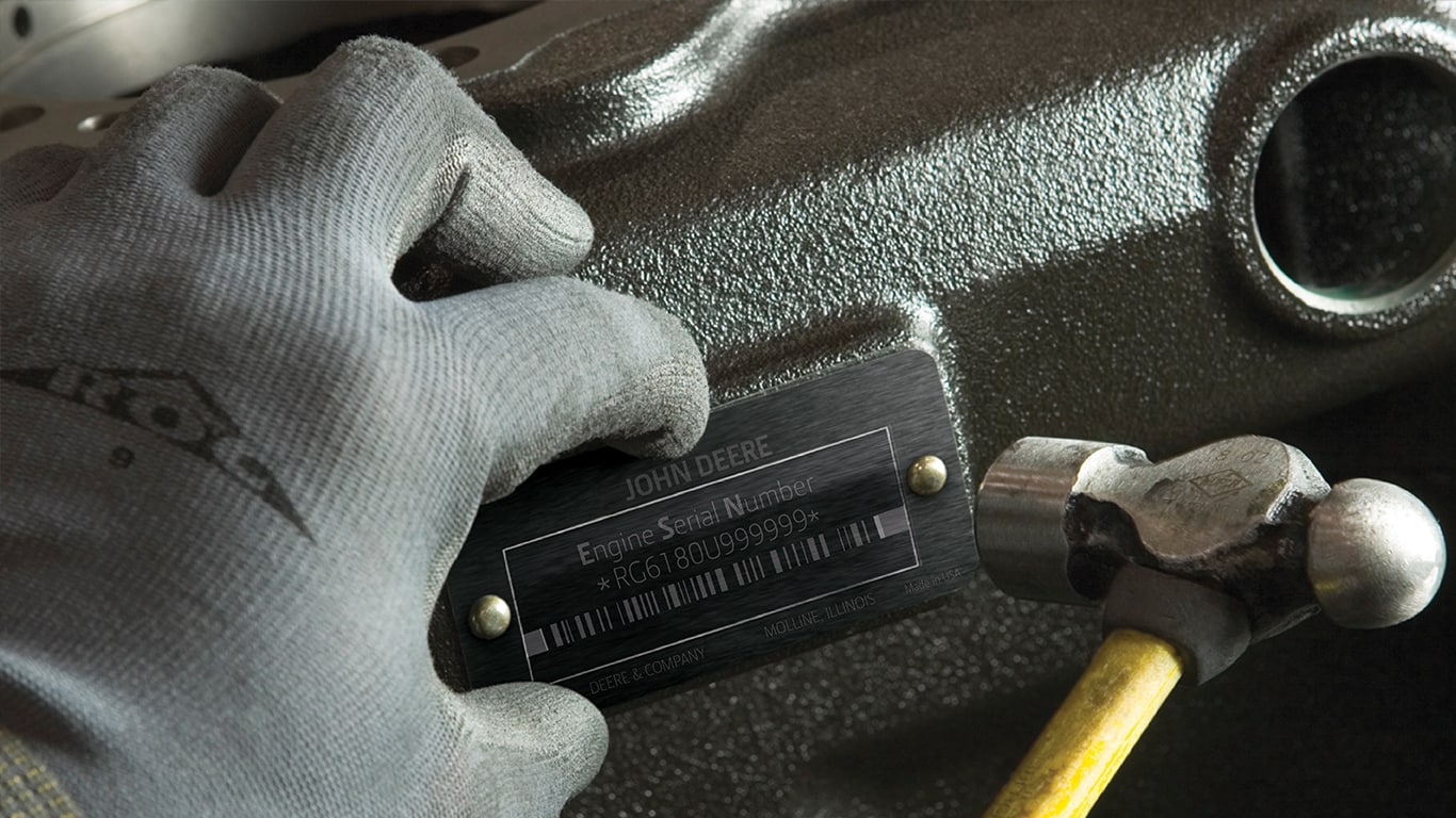 Una persona que lleva un guante gris sostiene un martillo mientras instala una placa de número de serie del motor John Deere en un motor