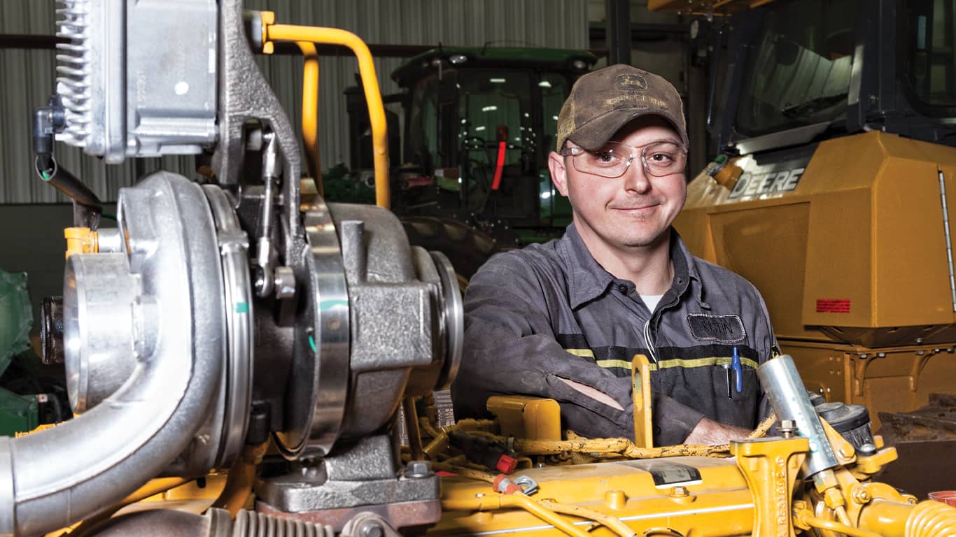 Un hombre con gafas de protección sonríe junto a un motor John Deere