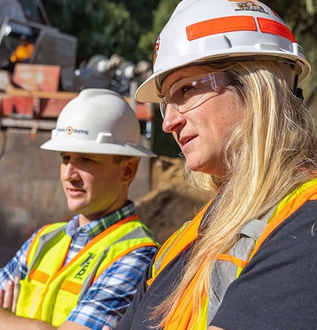 Un hombre y una mujer con cascos de obra y chalecos de seguridad miran un equipo de construcción