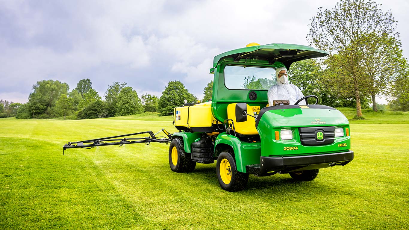 Un encargado del mantenimiento de espacios verdes con ropa de protección trabaja en el césped de un campo de golf con un pulverizador John Deere.