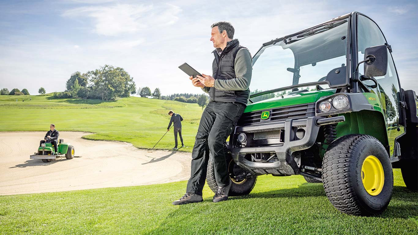 Un encargado jefe de mantenimiento revisa los datos en su tableta mientras dos trabajadores trabajan en un campo de golf en el fondo.