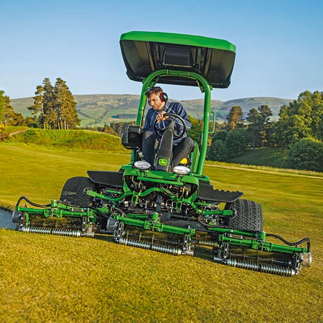 Un encargado de mantenimiento trabaja en una zona de césped con una segadora de greens John Deere vista desde el frente.