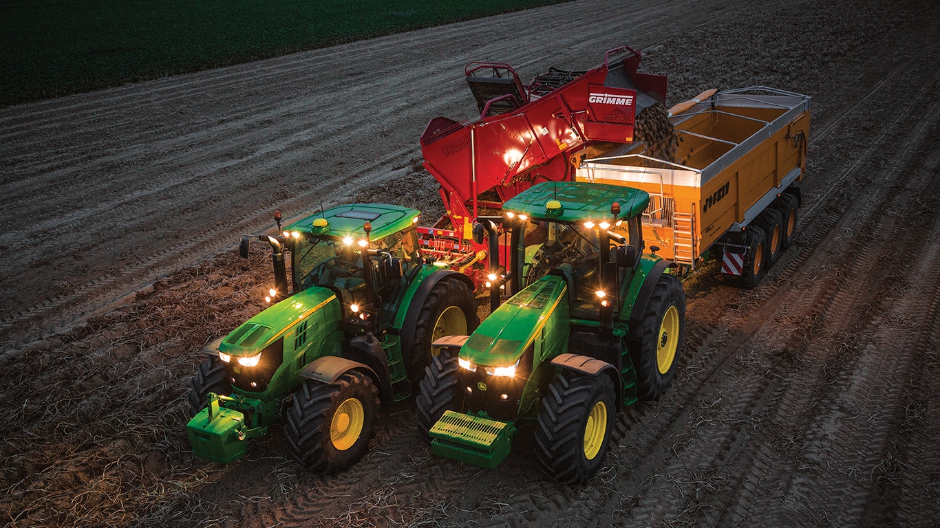 Un tractor que tira de una m&aacute;quina cosechadora de patatas por la noche est&aacute; descargando patatas al carro tirado por otro tractor