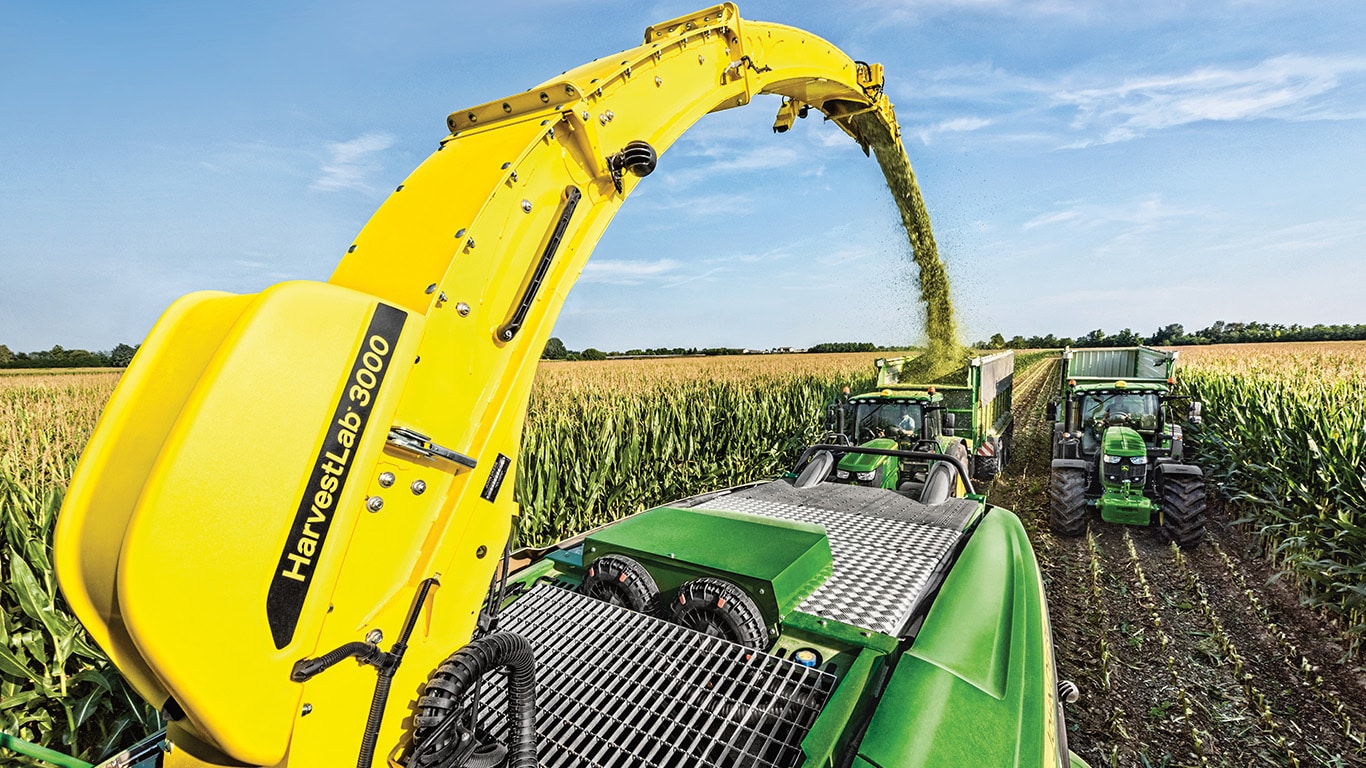 Vista de la parte trasera de la picadora de forraje autopropulsada de la serie 9000 John Deere con dos carros de ensilado tirados por tractores John Deere directamente detr&aacute;s